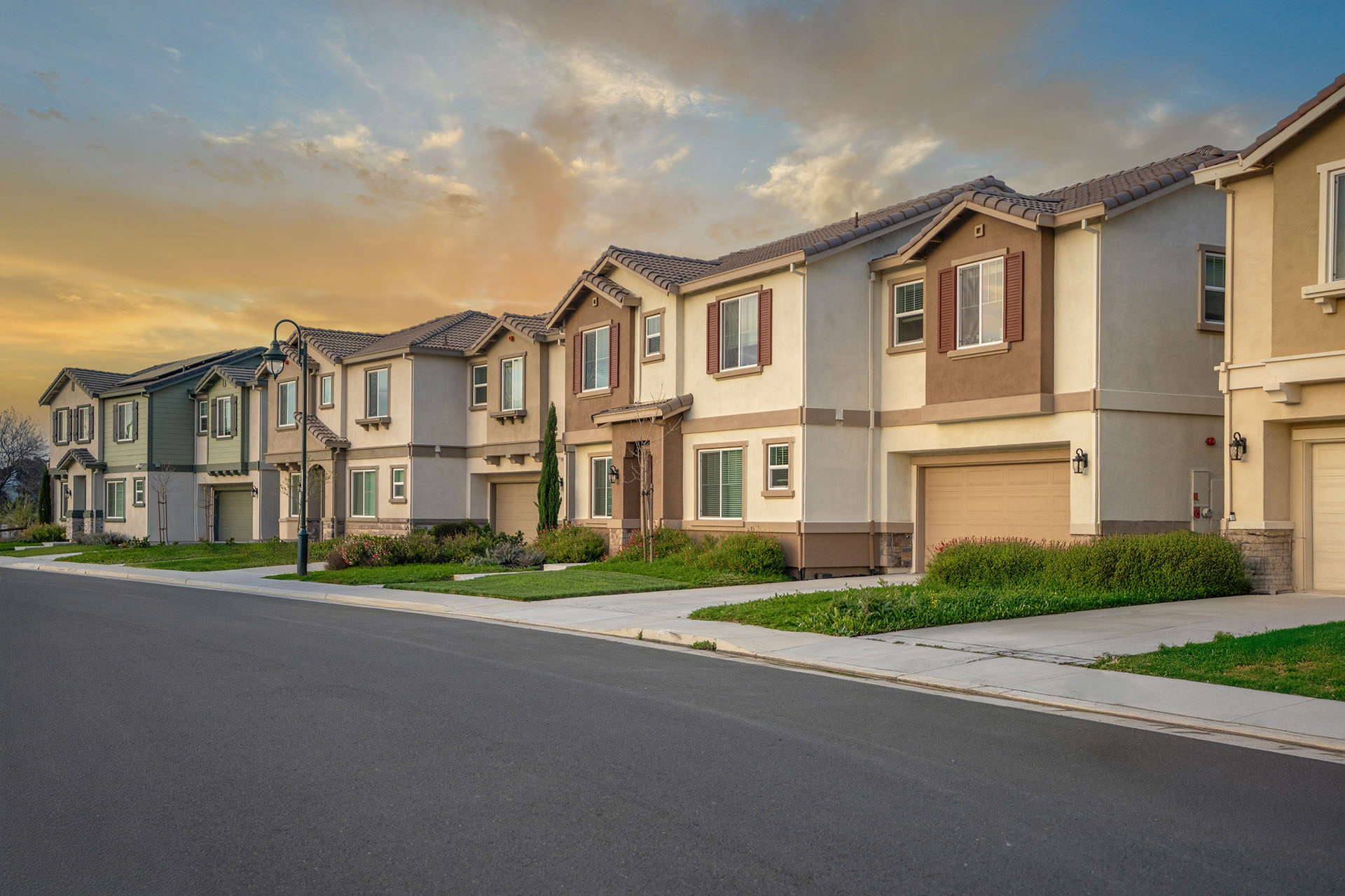 Row of newly constructed suburban townhouses with modern design, neutral exterior colors, and manicured landscaping along residential street at sunset
